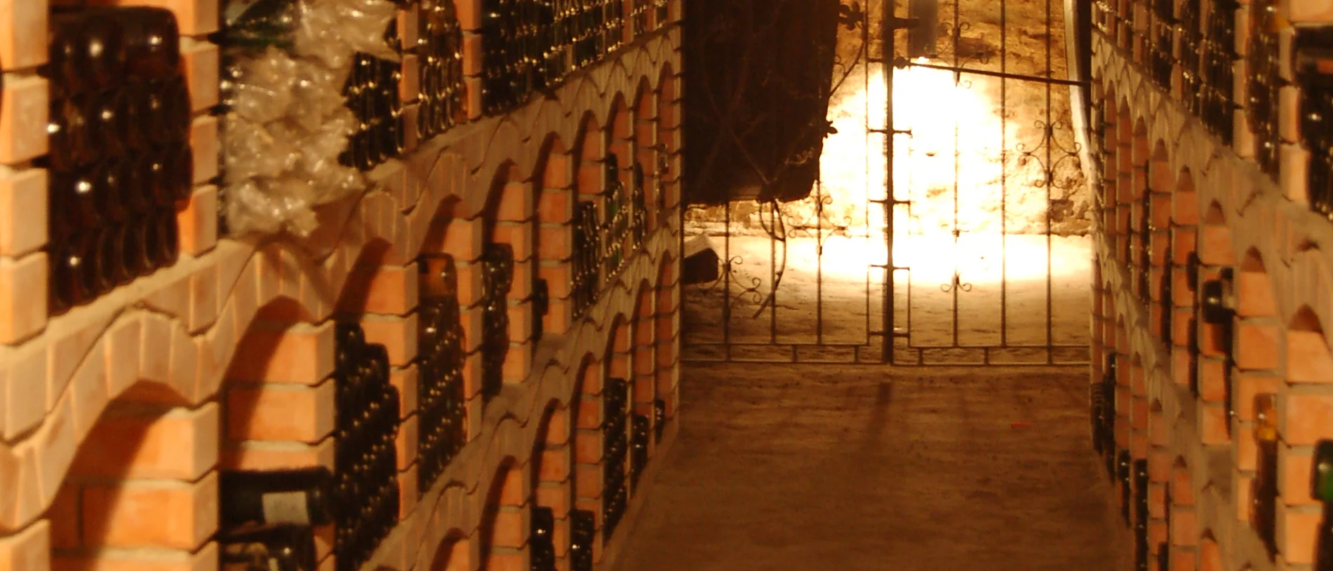 Une cave voûtée en brique : des centaines de bouteilles couchées dans des niches cintrées, vues par le fond, et une grille éclairée au bout de l'allée.